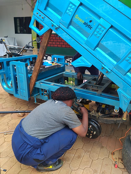 Person crouches to repair the rear wheel and suspension of a bright blue three-wheeled utility vehicle with its cargo bed tilted up.