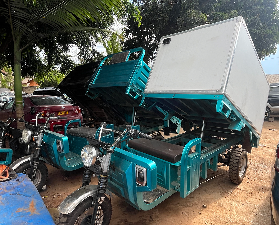 Two turquoise three-wheeled cargo motorcycles with their box beds tilted up, parked under trees in a lot.